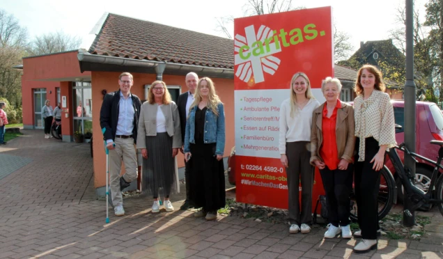 Gruppenfoto von sieben Personen vor einem roten Gebäude mit einem großen Caritas-Schild. Im Hintergrund sind weitere Personen und ein Fahrrad sichtbar.