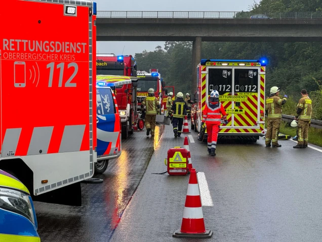 Einsatzfahrzeuge und Rettungskräfte auf einer nassen Straße, darunter Rettungswagen, Feuerwehrfahrzeuge und Personen in Einsatzkleidung. Verkehrshütchen sichern den Bereich ab, im Hintergrund ist eine Brücke sichtbar.