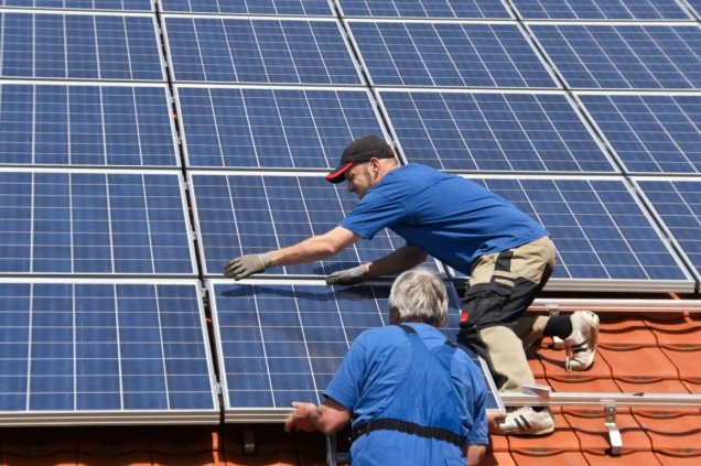 Zwei Personen installieren Solarmodule auf einem Ziegeldach. Eine Person kniet, die andere steht. Beide tragen blaue Arbeitskleidung und Handschuhe.