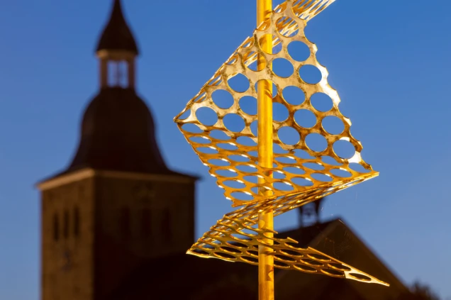 Eine goldene Skulptur aus gelochten Metallplatten vor einem unscharfen Hintergrund mit einem Kirchturm bei blauem Himmel.