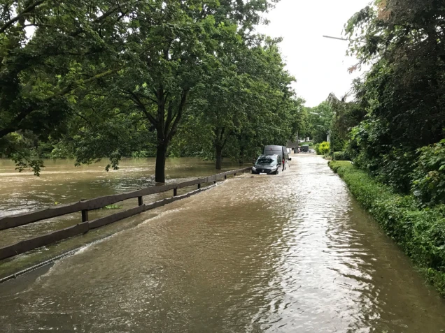 Eine überflutete Straße mit Autos, die im Wasser stehen. Auf der linken Seite sind Bäume und ein Holzzaun, rechts ist eine Hecke zu sehen.