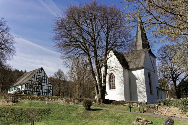 Eine weiße Kirche mit spitzem Turm und roten Fenstern steht neben einem Fachwerkhaus in einer ländlichen Umgebung mit Bäumen und Rasenflächen unter blauem Himmel.