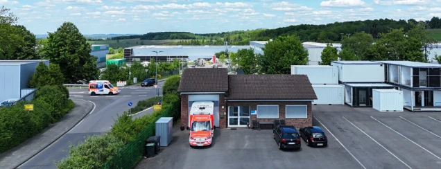 Eine Straße mit einem Rettungswagen, parkenden Autos und einem Gebäude im Vordergrund. Im Hintergrund befinden sich Bäume, Industriehallen und Container.