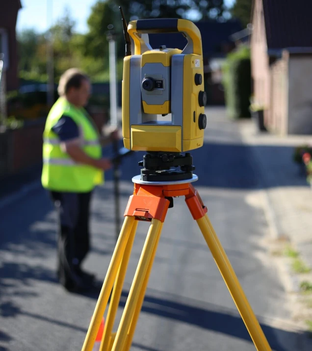 Eine gelb-graue Vermessungsstation auf einem gelben Stativ mit orangenen Elementen steht auf einer Straße. Im Hintergrund eine Person in gelber Warnweste unscharf sichtbar.
