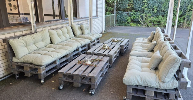 Eine Sitzgruppe aus Holzpaletten mit hellgrünen Polstern, bestehend aus zwei Sofas und zwei Tischen. Die Tische sind mit Kerzen und Dekoration verziert. Im Hintergrund ist eine Hauswand und eine Hecke zu sehen.