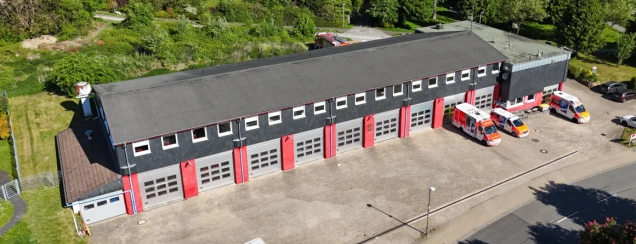 Luftaufnahme einer Feuerwehrwache mit grauem Dach, roter und grauer Fassade sowie mehreren Garagentoren. Vor dem Gebäude stehen Rettungsfahrzeuge und Autos. Im Hintergrund ist Grünfläche sichtbar.
