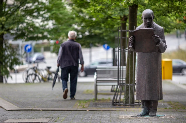 Bronzestatue eines Mannes, der eine Zeitung liest, steht auf einem Gehweg neben einem Baum. Im Hintergrund geht ein Mann mit Regenschirm vorbei, Fahrräder und Autos sind sichtbar.