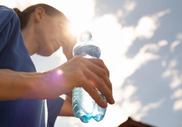 Eine Person in blauem Shirt hält eine Plastikflasche mit Wasser. Die Sonne scheint im Hintergrund, teilweise verdeckt durch Wolken, und erzeugt eine Lichtreflexion.