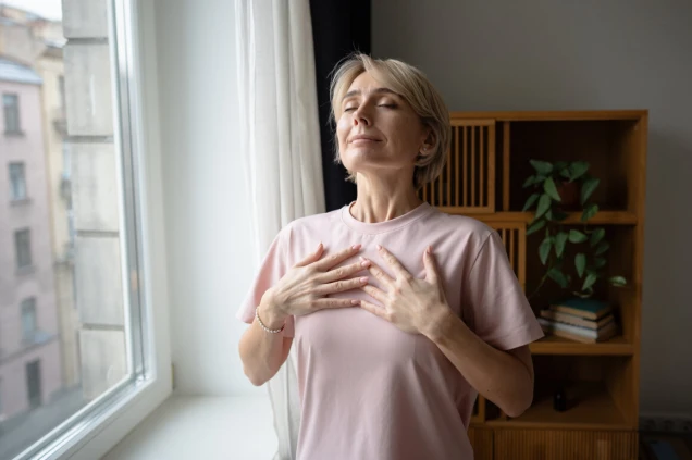 Eine Frau in einem rosa T-Shirt steht vor einem Fenster und hält beide Hände auf ihre Brust. Sie hat die Augen geschlossen und wirkt entspannt. Im Hintergrund ist ein Regal mit einer Pflanze und Büchern zu sehen.