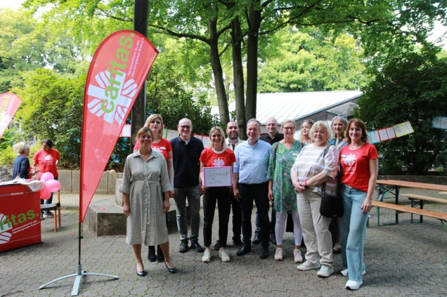 Eine Gruppe von Personen steht vor einem roten Caritas-Banner in einem Außenbereich mit Bäumen. Einige tragen rote T-Shirts mit dem Caritas-Logo.