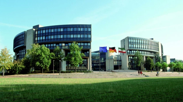 Landtagsgebäude Nordrhein-Westfalen in Düsseldorf. Moderne Gebäude mit Glasfassaden und runden Elementen, davor eine Grünfläche mit Bäumen. Drei Fahnen: EU, Deutschland und Nordrhein-Westfalen, wehen im Wind.