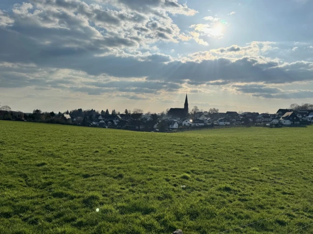 Grüne Wiese im Vordergrund, dahinter eine Siedlung mit Häusern und einer Kirche mit spitzem Turm. Der Himmel ist teils bewölkt mit Sonnenstrahlen.