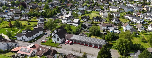 Luftaufnahme einer Wohnsiedlung mit vielen Einfamilienhäusern, Gärten und Grünflächen. Im Vordergrund ein Gebäude mit Solarpanels und roten Toren.