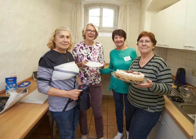 Vier Frauen stehen in einer Küche. Zwei Frauen halten einen Teller mit Kuchen. Im Hintergrund sind ein Fenster und Küchenmöbel zu sehen.