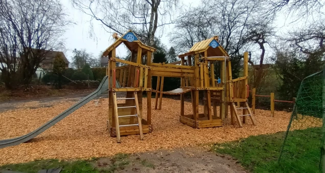 Ein hölzlicher Spielplatz mit zwei verbundenen Türmen, Rutschen und Kletterelementen, umgeben von Holzschnitzeln. Im Hintergrund stehen Bäume und Häuser.