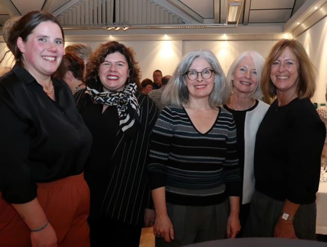 Fünf lächelnde Frauen stehen nebeneinander in einem Raum mit weißer Wand und moderner Deckenbeleuchtung. Im Hintergrund sind weitere Personen zu sehen.