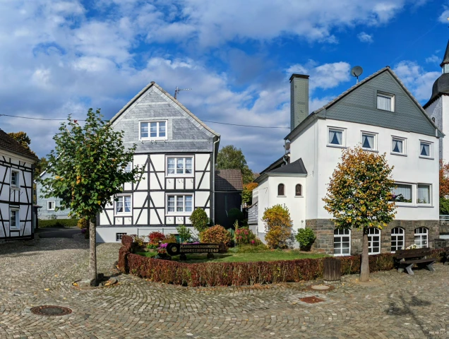 Ein Kopfsteinpflasterplatz mit Fachwerkhäusern, einer Kirche mit Turm und Spitzdach sowie Bäumen im herbstlichen Laub. Im Vordergrund stehen Sitzbänke.