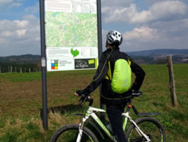 Eine Person mit Fahrradhelm und gelbem Rucksack steht neben einem weißen Fahrrad und betrachtet eine Knotenpunkttafel mit Informationen zum Radverkehr in einer ländlichen Landschaft.