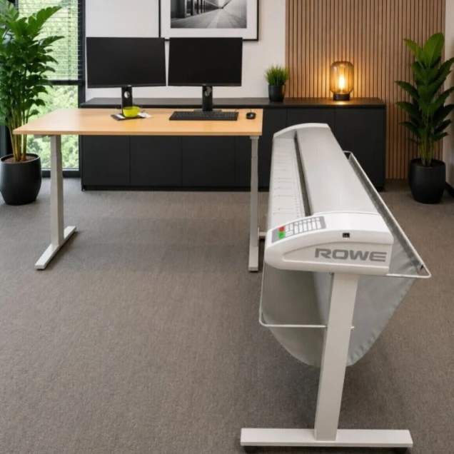 Modern eingerichtetes Büro mit einem höhenverstellbaren Schreibtisch, zwei Monitoren, einer Tastatur und einer Maus. Im Vordergrund ein großes Druckgerät mit der Aufschrift 'ROWE'. Im Hintergrund eine schwarze Kommode mit Dekoration, darunter eine Lampe und Pflanzen. Die Wand ist teils mit Holzpaneelen verkleidet, teils weiß, mit einem gerahmten Schwarz-Weiß-Foto. Fenster mit Jalousien und Ausblick auf grüne Bäume.