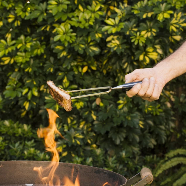 Eine Hand hält eine Grillzange mit einem Stück Fleisch über einen runden Metallgrill mit offener Flamme. Im Hintergrund sind grüne Büsche sichtbar.