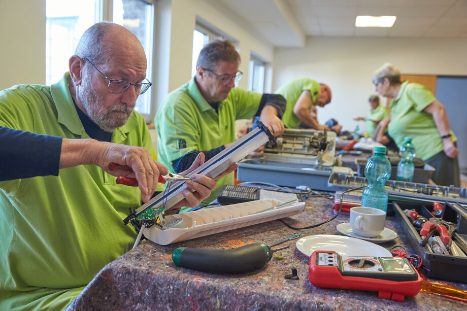 Mehrere Personen in grünen Polohemden reparieren elektronische Geräte an einem Tisch in einem Innenraum. Auf dem Tisch liegen Werkzeuge und Bauteile.