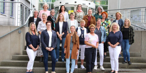 Eine Gruppe von Frauen steht auf einer Treppe vor einem modernen Gebäude mit Glasfront. Sie tragen verschiedene Freizeitkleidungen und lächeln in die Kamera.