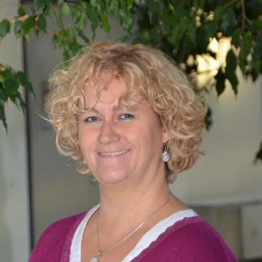 Eine Frau mit lockigem blondem Haar lächelt in die Kamera. Sie trägt ein lila Oberteil mit weißem Ausschnitt und steht vor einem unscharfen Hintergrund mit grünen Pflanzen.