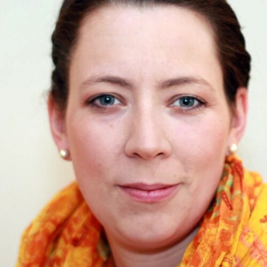Porträt einer Frau mit zurückgebundenem Haar, die direkt in die Kamera schaut. Sie trägt Ohrringe und einen bunten, orange-gelben Schal.
