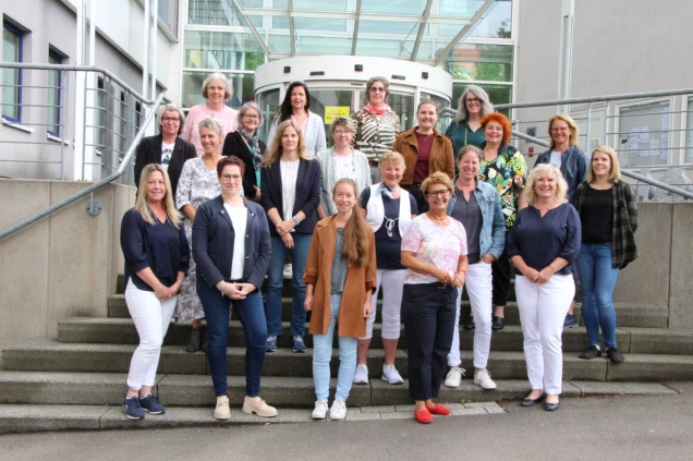 Eine Gruppe von Frauen steht auf einer Treppe vor einem modernen Gebäude mit Glasfront. Sie tragen verschiedene Freizeitkleidungen und lächeln in die Kamera.