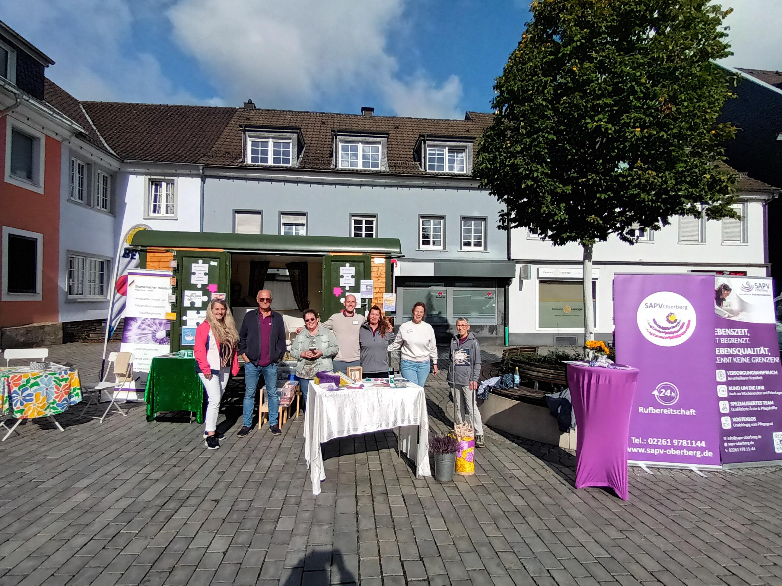 Eine Gruppe von Menschen steht auf einem Platz vor einem kleinen grünen Verkaufsstand. Links und rechts sind Tische mit Informationsmaterial und ein lila Banner.