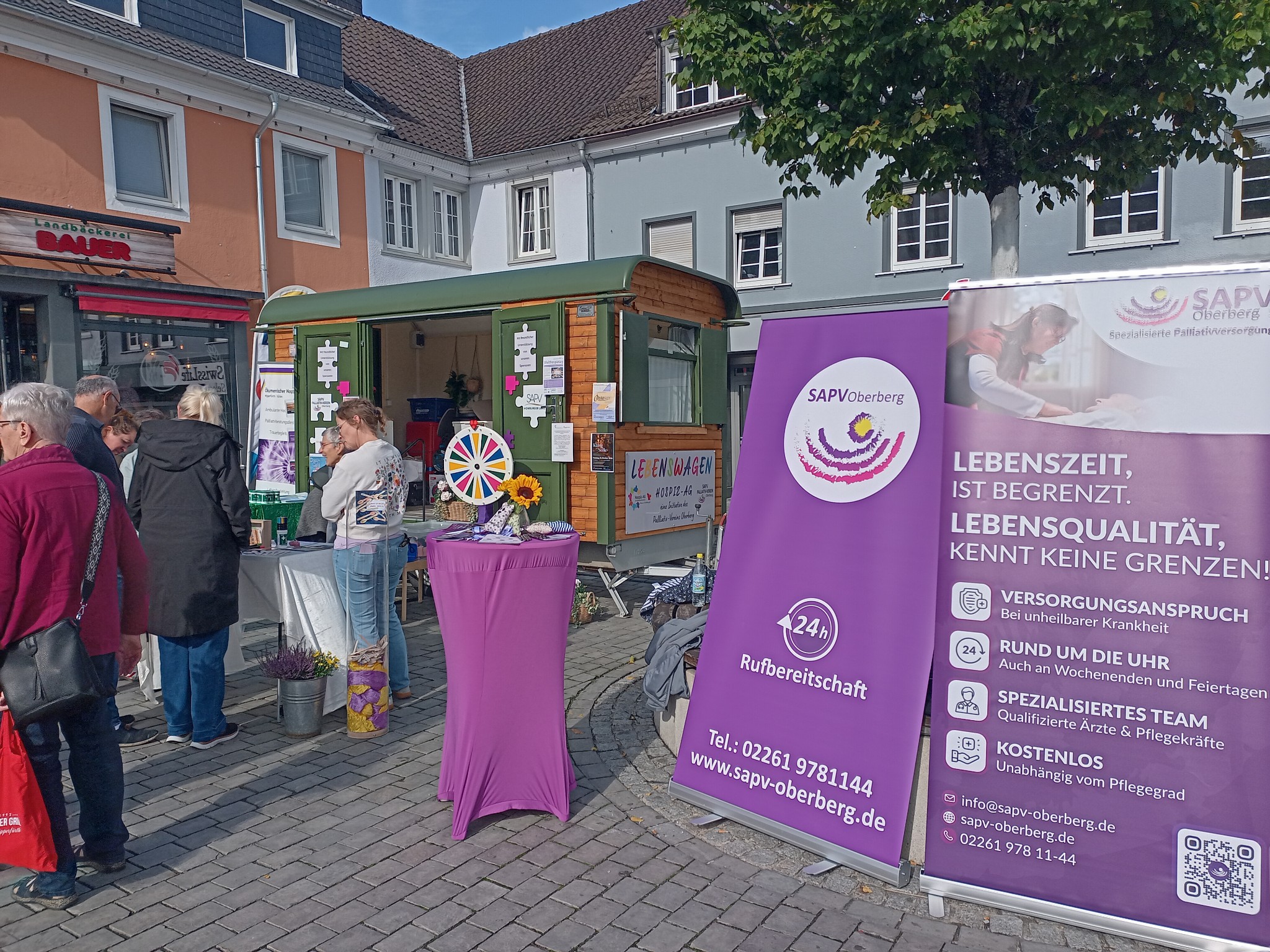 Menschen stehen an einem Stand mit Informationsmaterial in einer Fußgängerzone. Ein violetter Infostand von SAPV Oberberg und ein kleiner Verkaufswagen sind zu sehen.