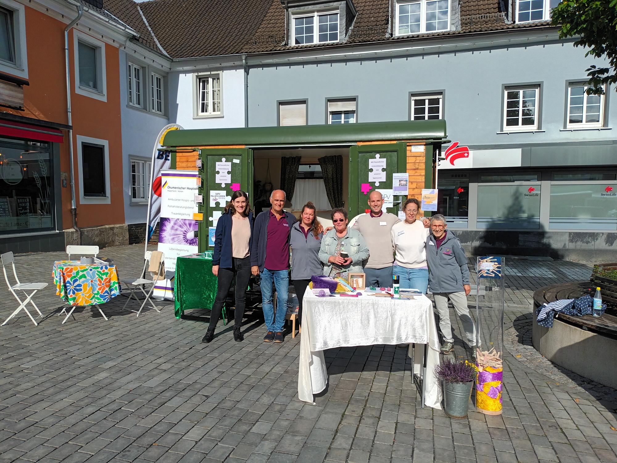 Eine Gruppe von Menschen steht vor einem kleinen Stand auf einem gepflasterten Platz. Der Stand hat grüne und holzfarbene Elemente, mit Flyern und Dekoration. Neben dem Stand gibt es einen Tisch mit buntem Tischtuch und weißen Stühlen. Im Hintergrund sind Gebäude mit Fenstern und Schaufenstern sichtbar.