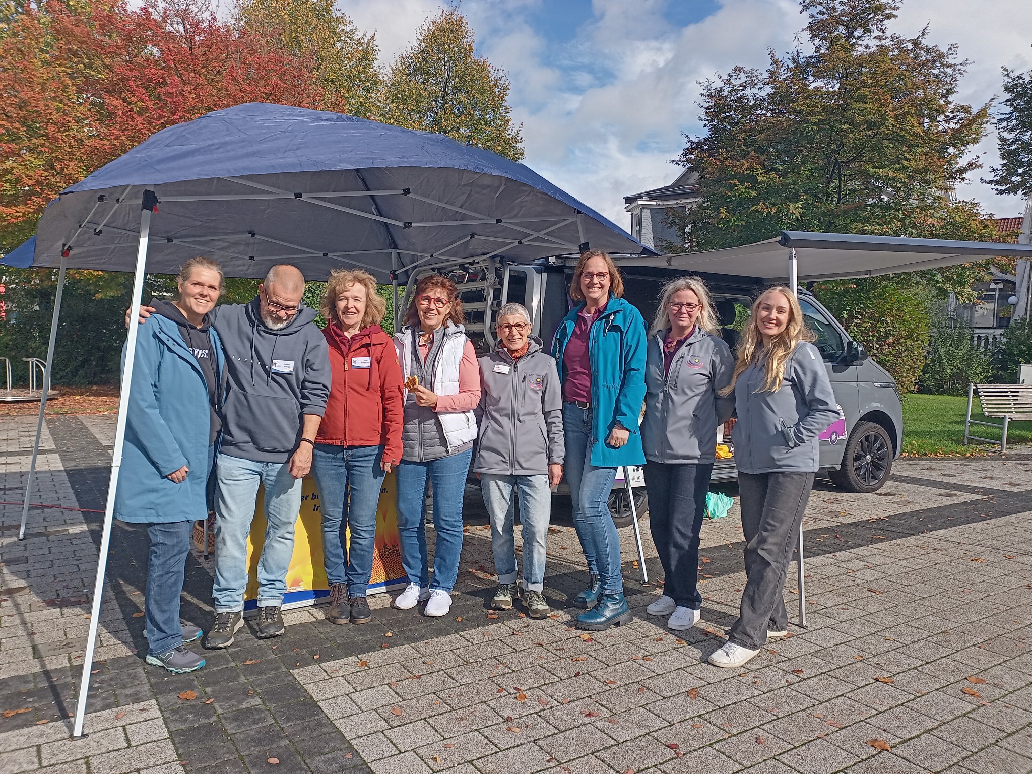 Gruppe von acht Personen steht unter einem Pavillon neben einem grauen Transporter. Im Hintergrund sind Herbstbäume und ein gepflasterter Platz sichtbar.
