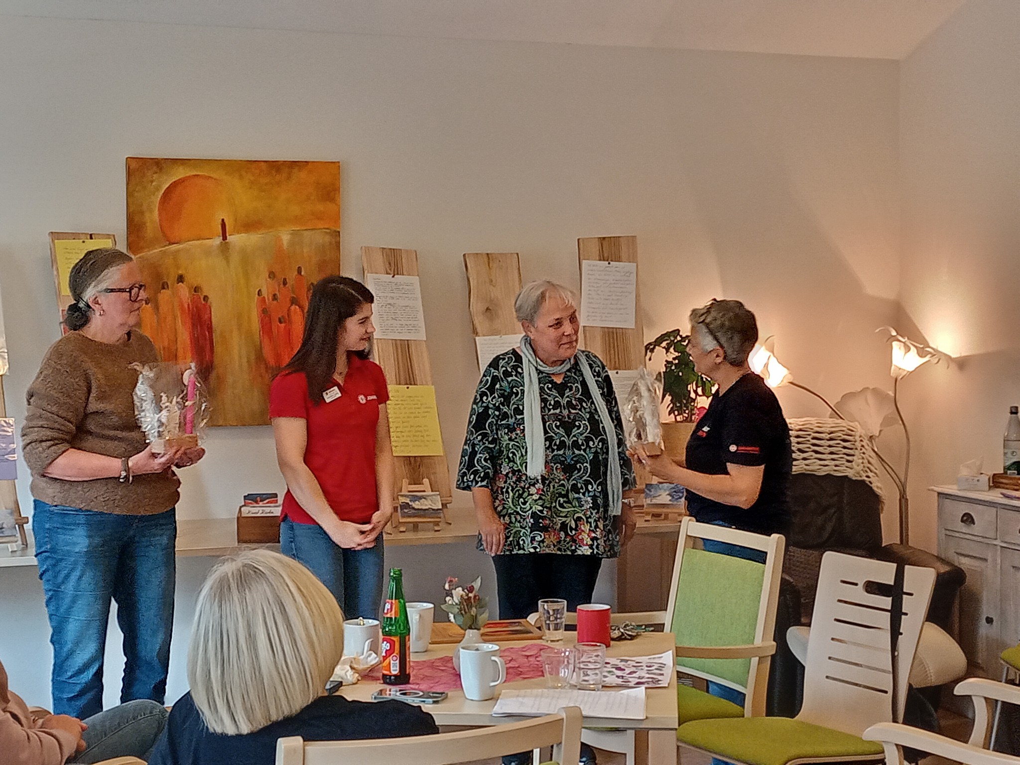 Vier Personen stehen in einem Raum, drei von ihnen im Gespräch. Zwei halten Geschenke in der Hand. Im Hintergrund sind Dekorationen und Möbel zu sehen.