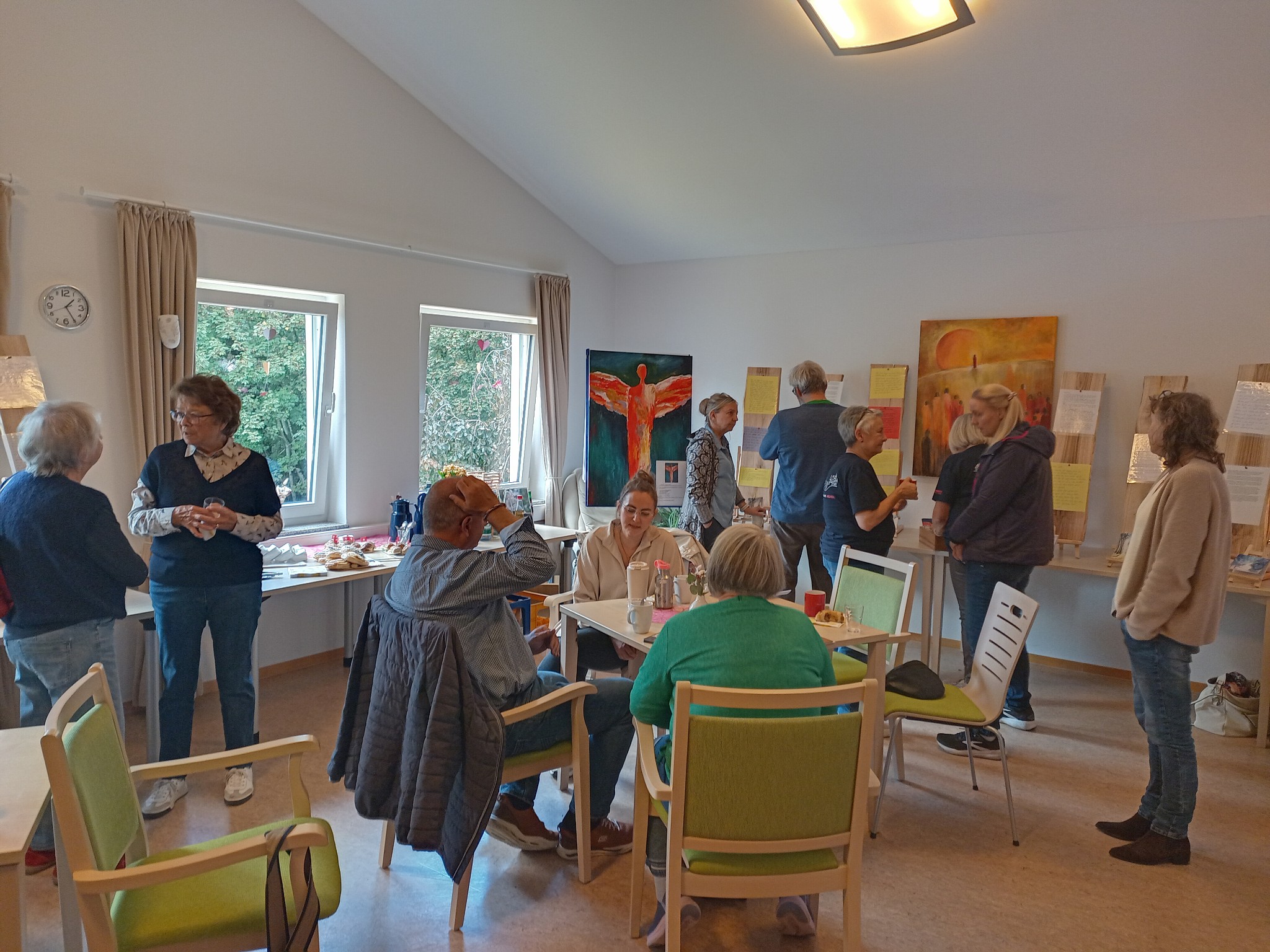 Mehrere Menschen stehen und sitzen in einem hellen Raum mit großen Fenstern. An einer Wand sind Gemälde und Texte auf Holztafeln ausgestellt.