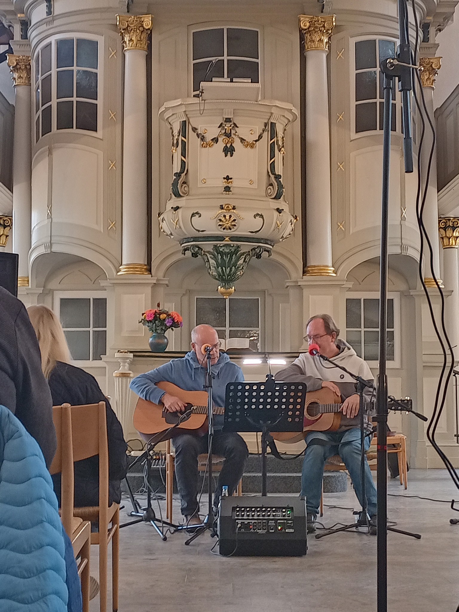 Zwei Personen sitzen in einer Kirche und spielen Akustikgitarren. Vor ihnen stehen Mikrofone und ein Notenständer. Der Kirchenraum ist mit goldenen Verzierungen dekoriert.