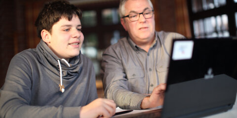 Ein jüngerer und ein älterer Mann sitzen an einem Tisch und schauen gemeinsam auf einen Laptop. Der Raum im Hintergrund hat Fenster und Backsteinwände.