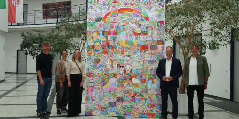 Fünf Personen stehen in einem modernen Gebäude vor einem großflächigen bunten Kunstwerk mit Motiven wie Regenbögen, Blumen, Friedenstauben und Gesichtern.