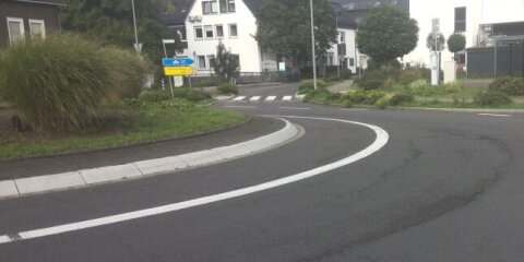Eine asphaltierte Straße mit einer Kurve, Straßenmarkierungen und einem begrünten Mittelstreifen. Im Hintergrund Gebäude, Schilder und ein Parkplatz sichtbar.