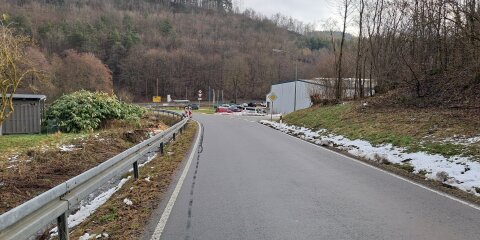 Eine asphaltierte Straße mit Leitplanke führt bergab in eine Kurve. Links und rechts sind schneebedeckte Grasflächen und Bäume. Im Hintergrund stehen Autos und ein Gebäude vor einem bewaldeten Hügel.