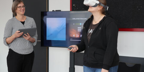 Eine Frau mit Brille hält ein Tablet und lächelt, während eine andere Frau eine VR-Brille trägt und einen Controller hält. Im Hintergrund ist ein Bildschirm mit einer Unterwasseraufnahme und der Schrift 'Delfintraum' zu sehen.