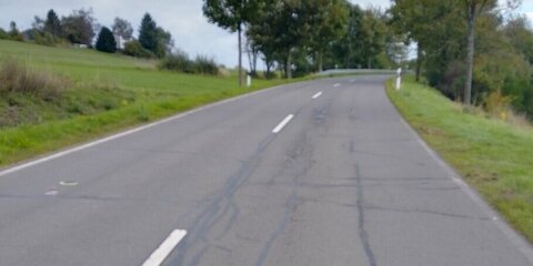 Eine asphaltierte Landstraße mit weißen Markierungen und Reparaturspuren, die durch eine grüne Landschaft mit vereinzelten Bäumen und Häusern führt. Der Himmel ist bewölkt.