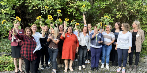 Eine Gruppe von Frauen steht auf gepflastertem Boden vor einer grünen Hecke. Sie halten Sonnenblumen in die Luft und lächeln in die Kamera.