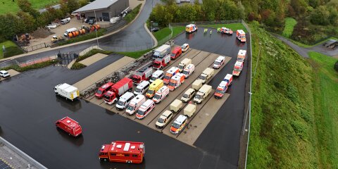 Drohnenaufnahme eines Parkplatzes mit einer Vielzahl von Einsatzfahrzeugen, darunter Rettungswagen, Feuerwehrfahrzeuge und andere Einsatzfahrzeuge. Im Hintergrund ein Gebäude, Grünflächen und Straßen.