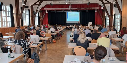 Ein großer Raum mit vielen Menschen, die an Tischen sitzen und auf eine Bühne mit einem Vortragenden und einer Präsentation blicken. Der Raum hat Holzsäulen und große Fenster.