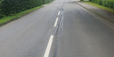 Straße mit Mittelstreifen, teilweise mit dunklen Streifen auf dem Asphalt. Links und rechts sind Gehwege, Hecken und Bäume. Im Hintergrund eine leichte Steigung.
