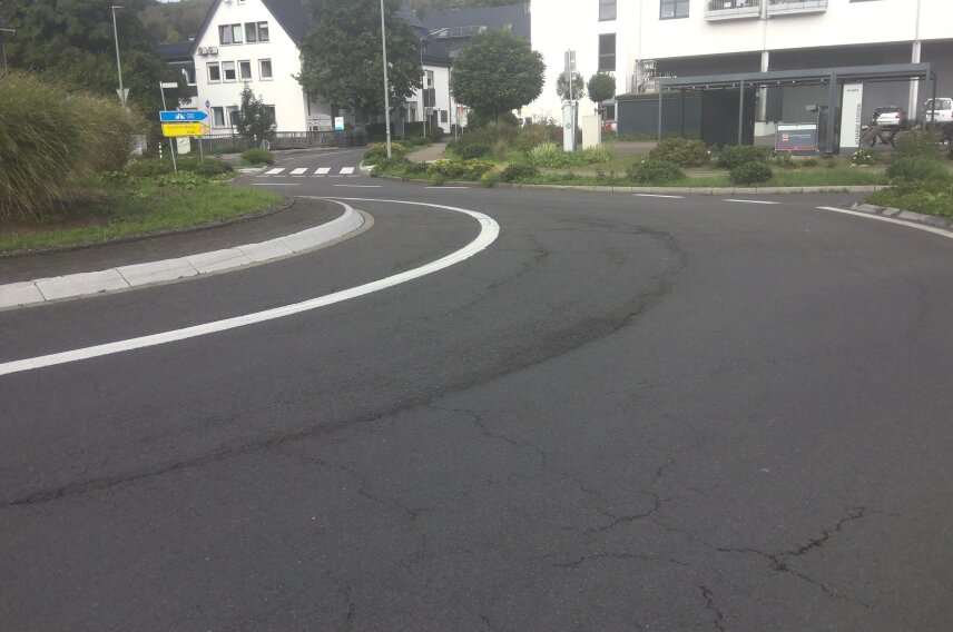 Eine asphaltierte Straße mit einer Kurve, Straßenmarkierungen und einem begrünten Mittelstreifen. Im Hintergrund Gebäude, Schilder und ein Parkplatz sichtbar.