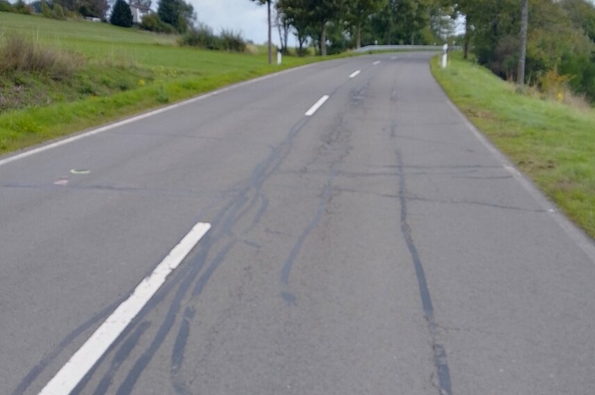 Eine asphaltierte Landstraße mit weißen Markierungen und Reparaturspuren, die durch eine grüne Landschaft mit vereinzelten Bäumen und Häusern führt. Der Himmel ist bewölkt.