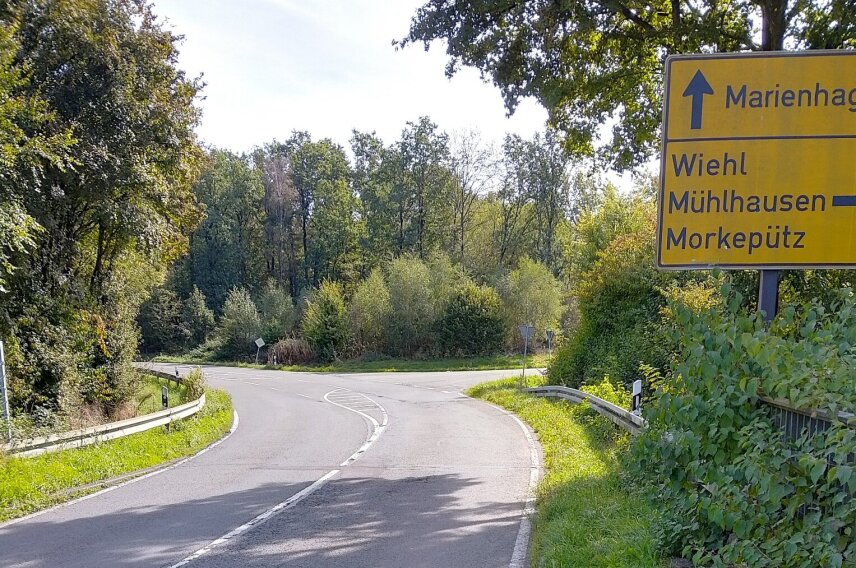 Straßenkreuzung mit Beschilderung. Gelbes Schild zeigt Richtungen nach Marienhagen, Wiehl, Mühlhausen und Morkepütz. Umgeben von grüner Vegetation und Bäumen.