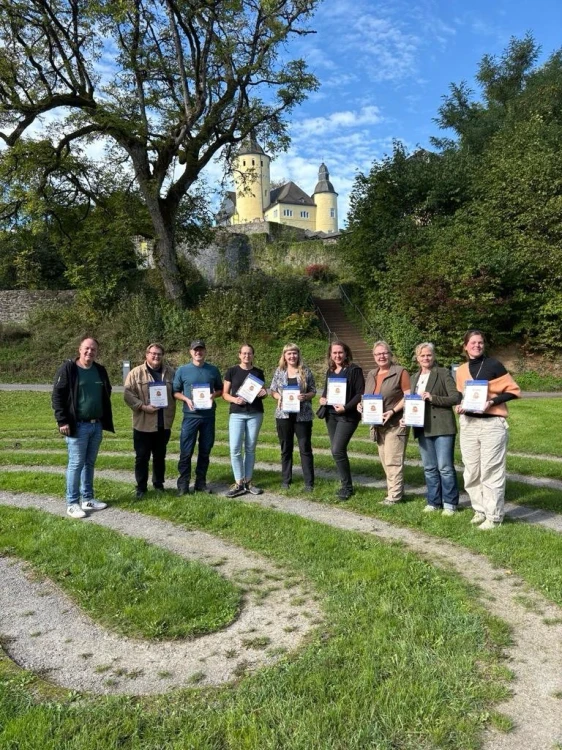 Neun Personen stehen auf einer Wiese vor einem Schloss mit gelben Türmen. Sie halten Urkunden in den Händen. Im Hintergrund Bäume und eine Treppe.
